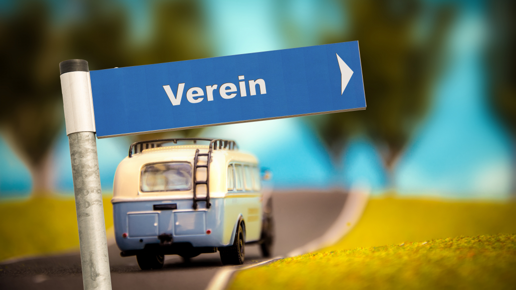 Bild mit einem Bus in Hintergrund auf einer Straße. Im Vordergrund ein Schild mit der Aufschrift "Verein" und einem Pfeil nach rechts